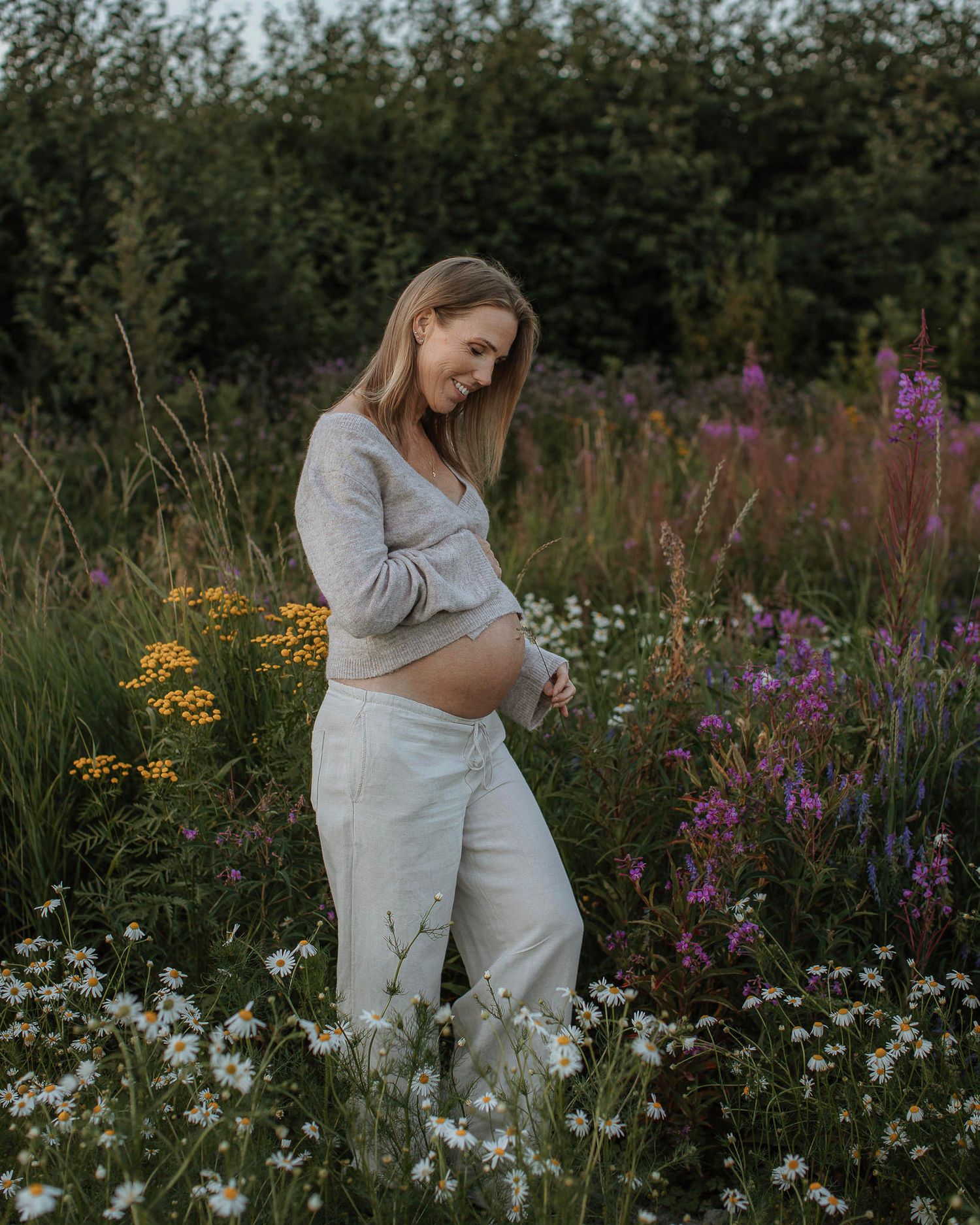 En gravid kvinna står på en sommaräng med blomstrande blommor. Hon håller i en kvist i ena handen och andra handen ovanför gravidmagen.