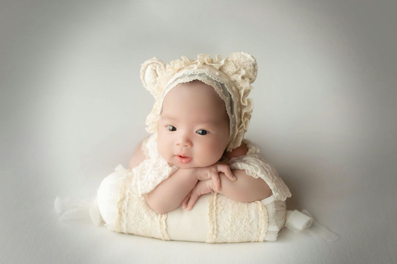 Newborn baby wearing cream-colored bonnet lying on soft white blanket in professional studio setting.