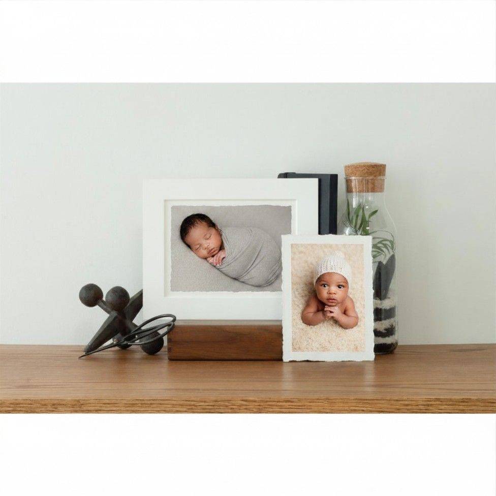 Two framed newborn baby photos displayed on a wooden shelf with decorative objects and a small terrarium.