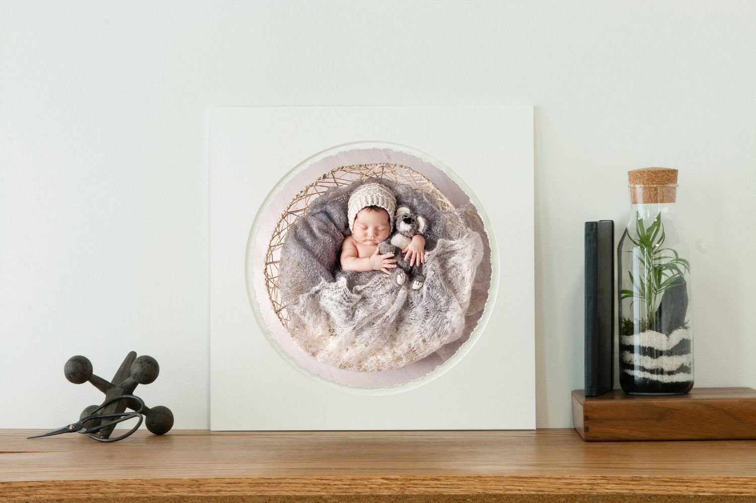 Newborn baby sleeping in a round basket on a white canvas print displayed on a wooden shelf with decorative items.