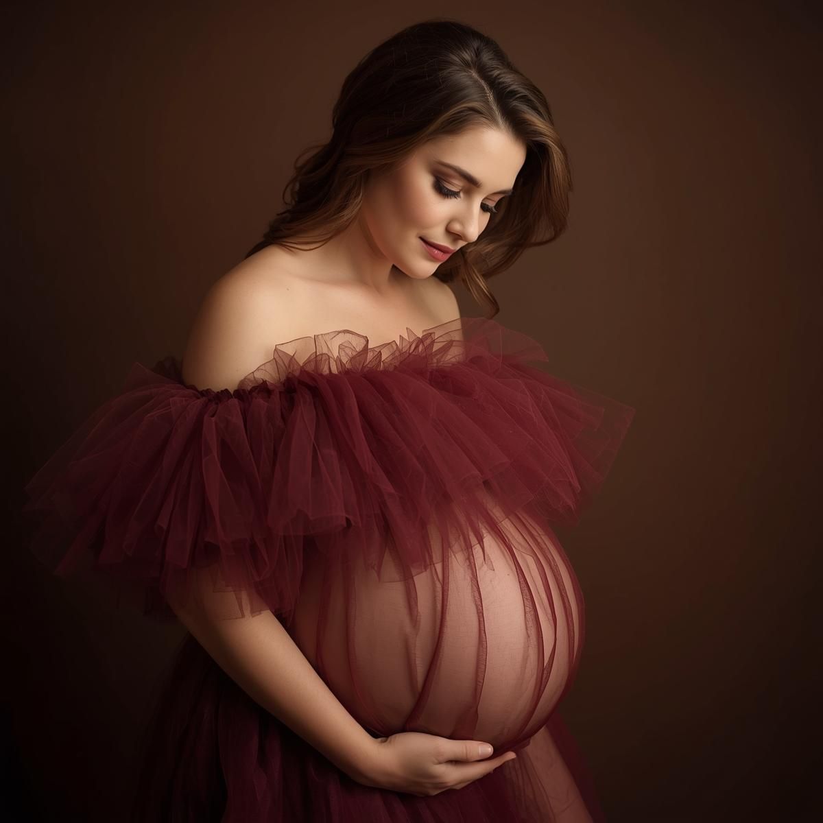 Maternity portrait in burgundy tulle gown with soft lighting against dark brown background.