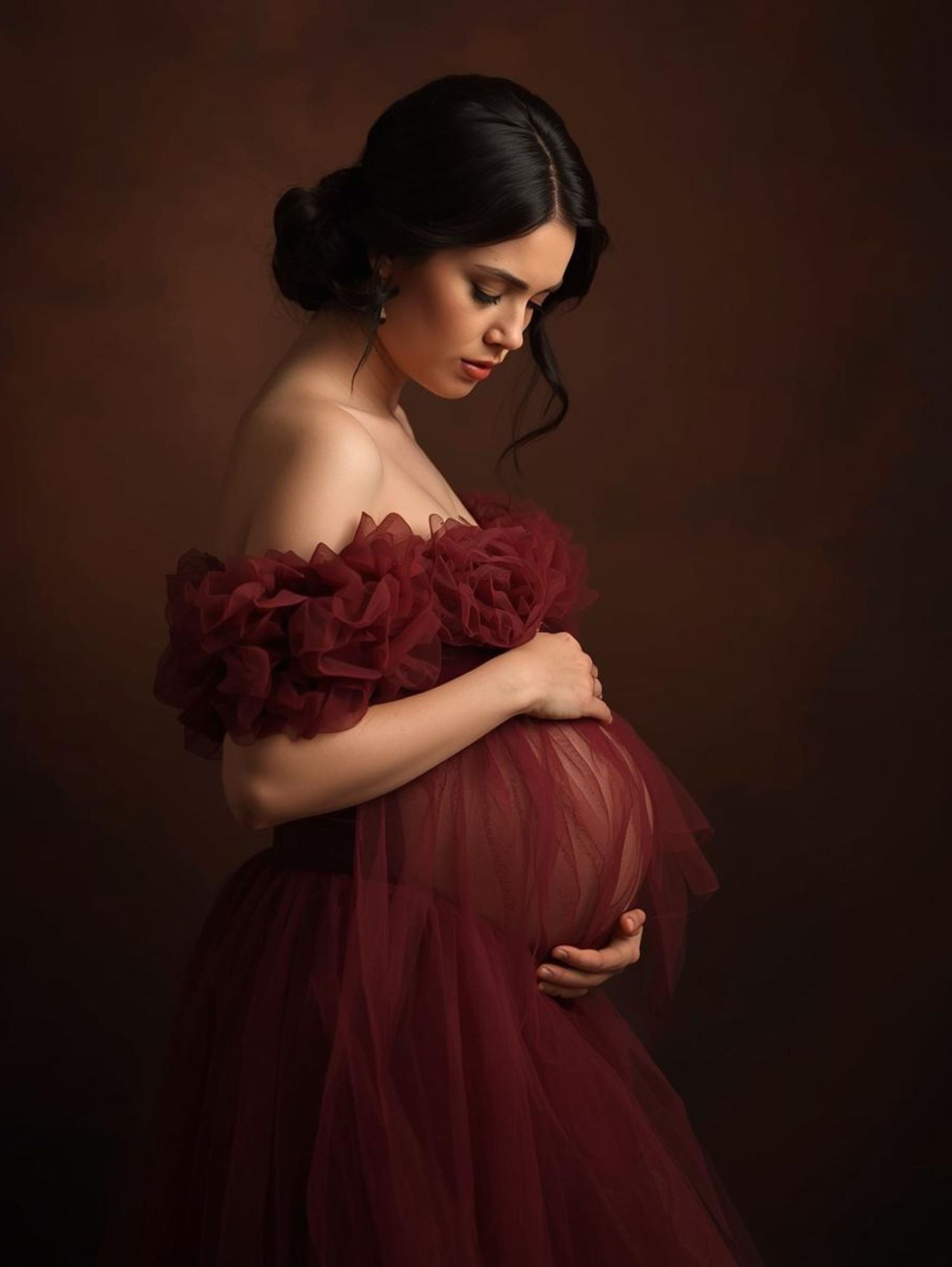 Artistic maternity portrait showing an expectant mother in a flowing burgundy tulle gown against a dark brown backdrop.