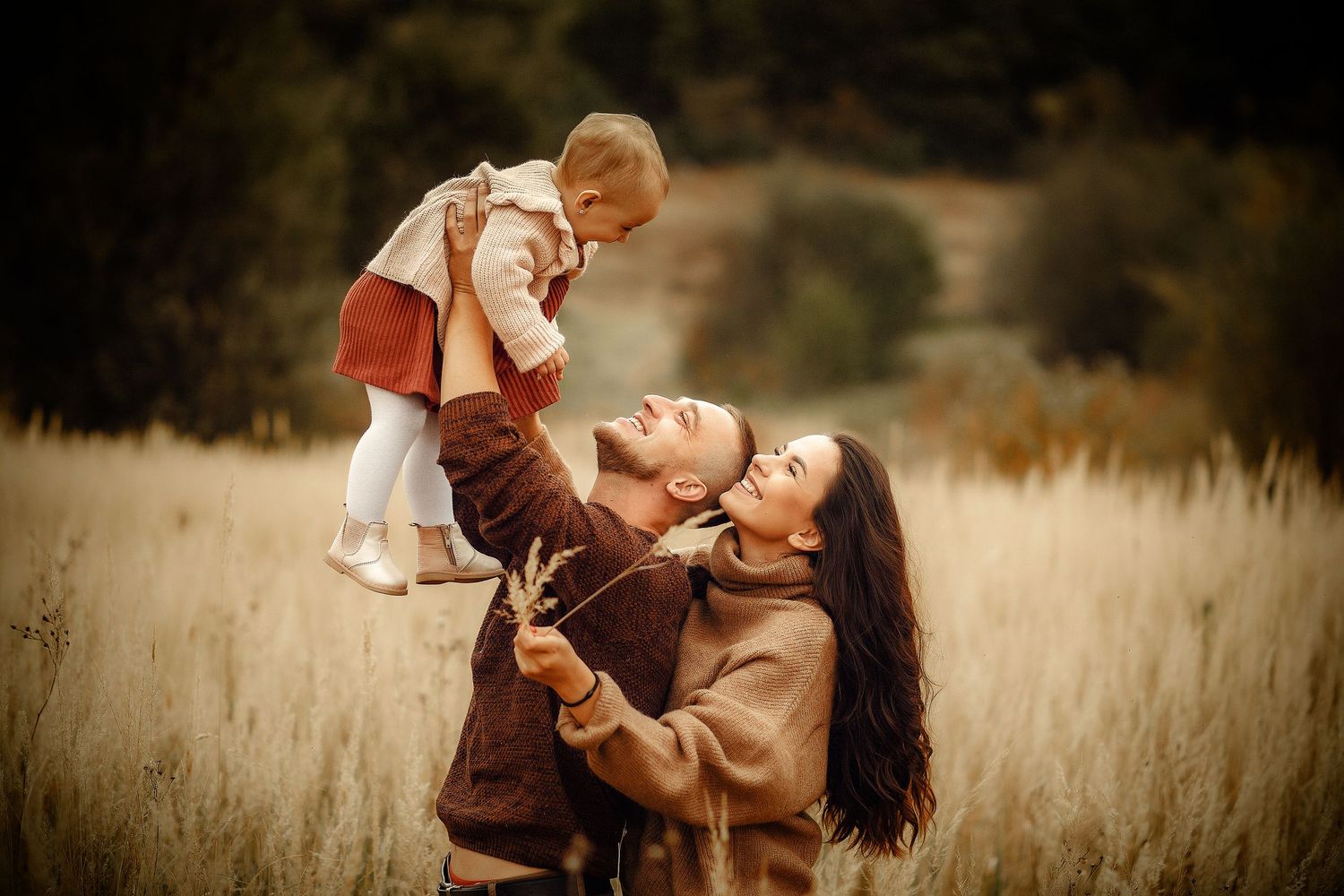 A joyful family in an autumn field, parents lifting their baby girl in the air, surrounded by golden grass.