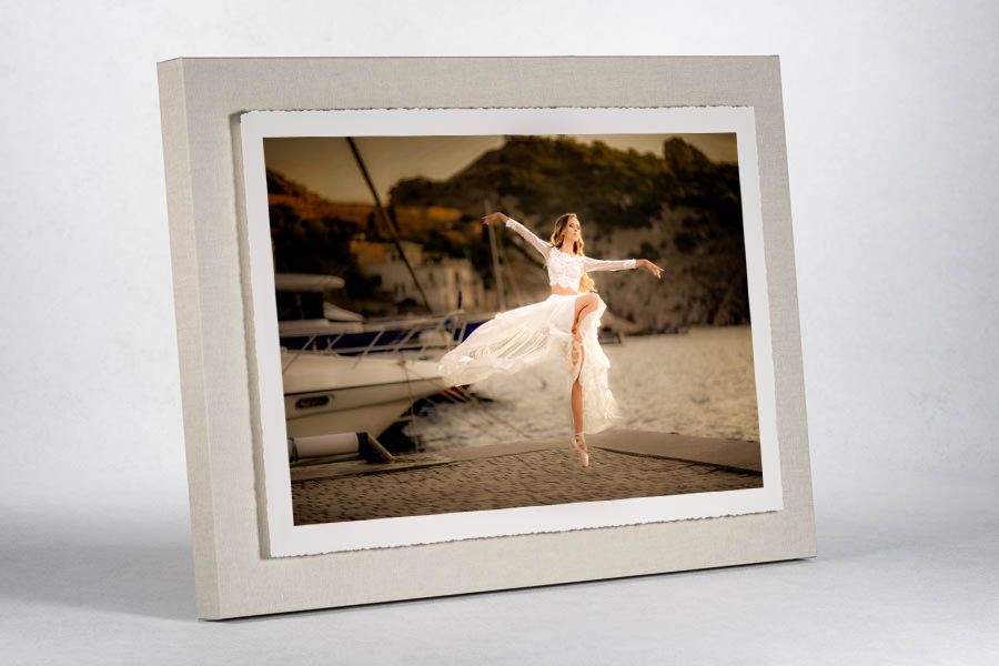 Framed photo of a ballet dancer in a white dress performing on a beach near a boat, displayed in a white picture frame.