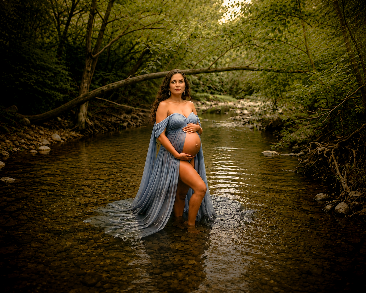Pregnant woman in blue flowing gown standing in a serene forest creek surrounded by lush green trees.