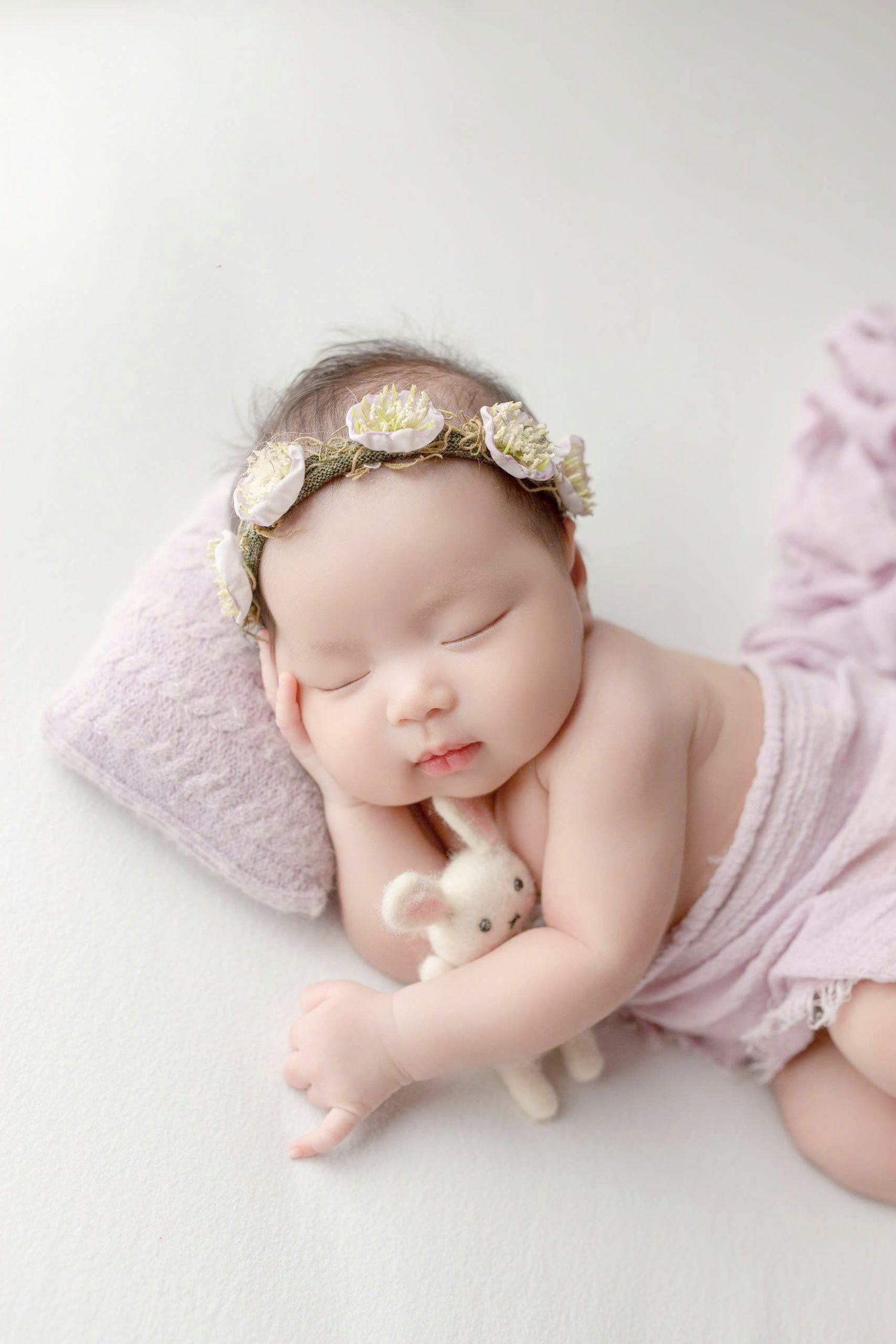 Sleeping newborn in pink outfit wearing delicate floral crown poses peacefully on white backdrop during professional photoshoot.