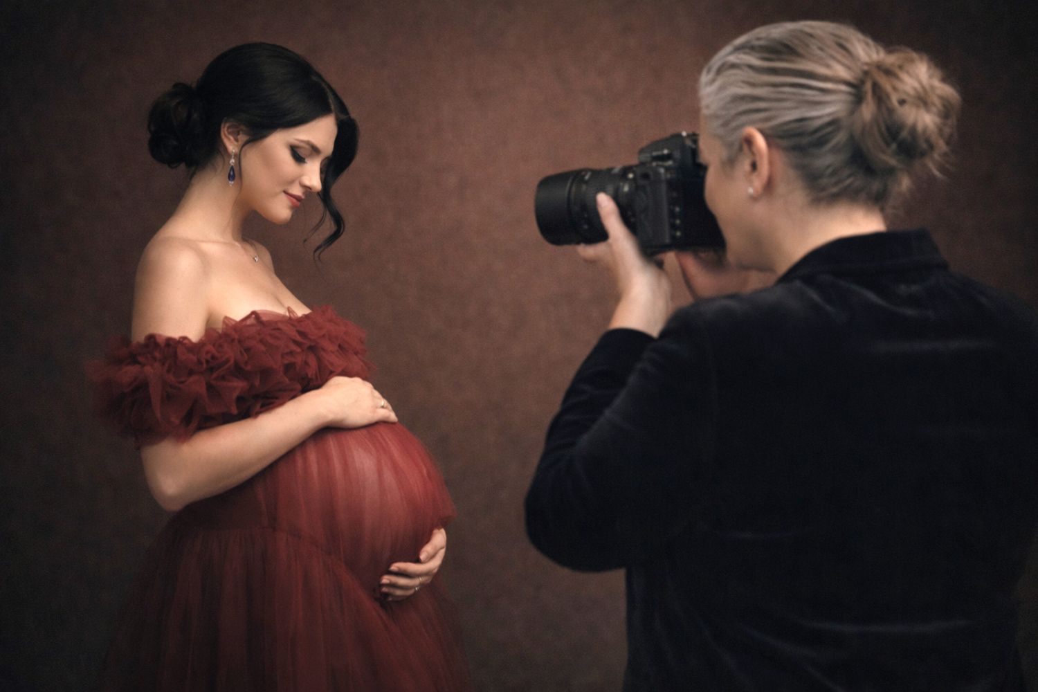 A photographer captures a maternity portrait of a woman wearing a flowing red off-shoulder dress against a dark backdrop.