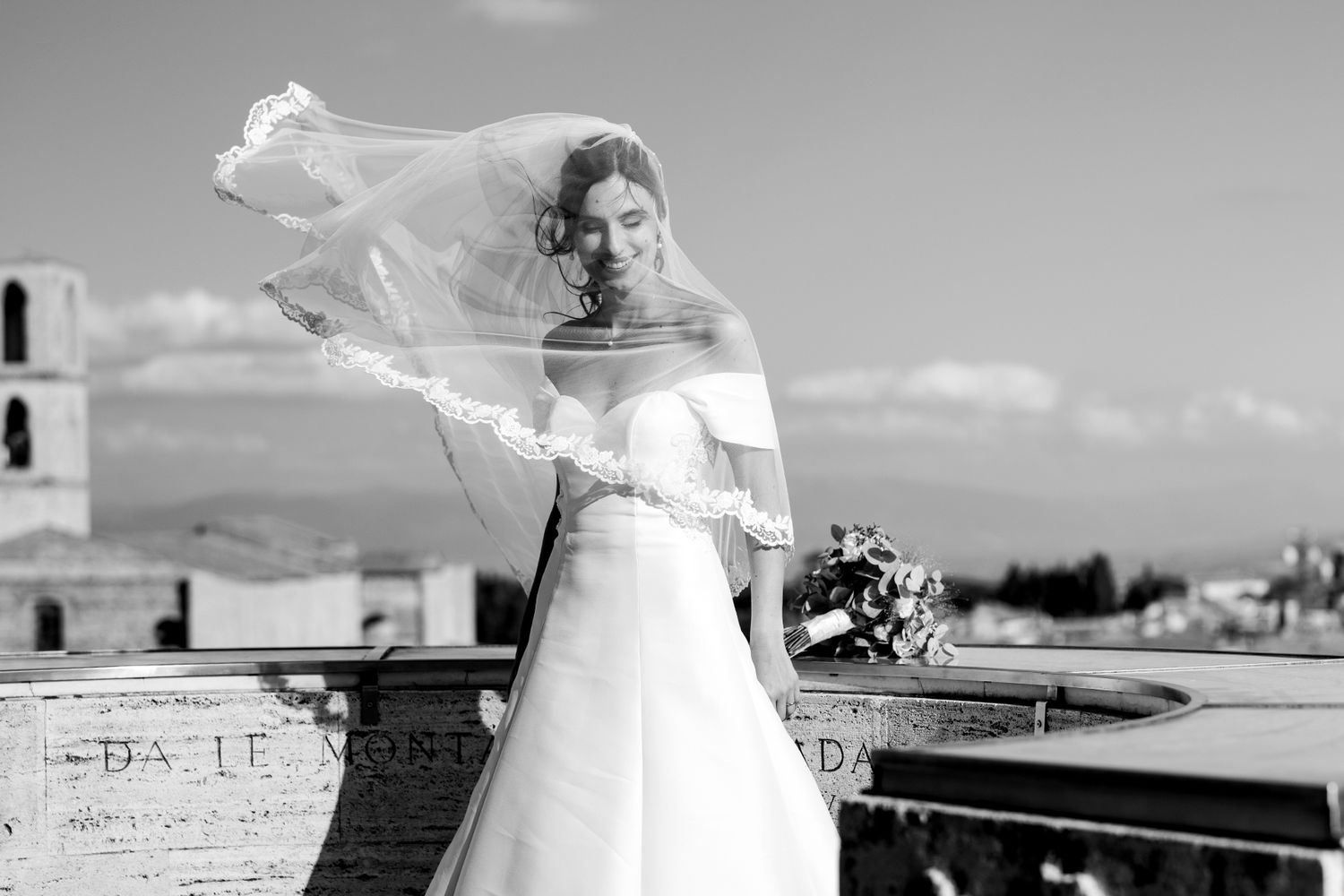 Black and white portrait of bride with windswept veil against city skyline.