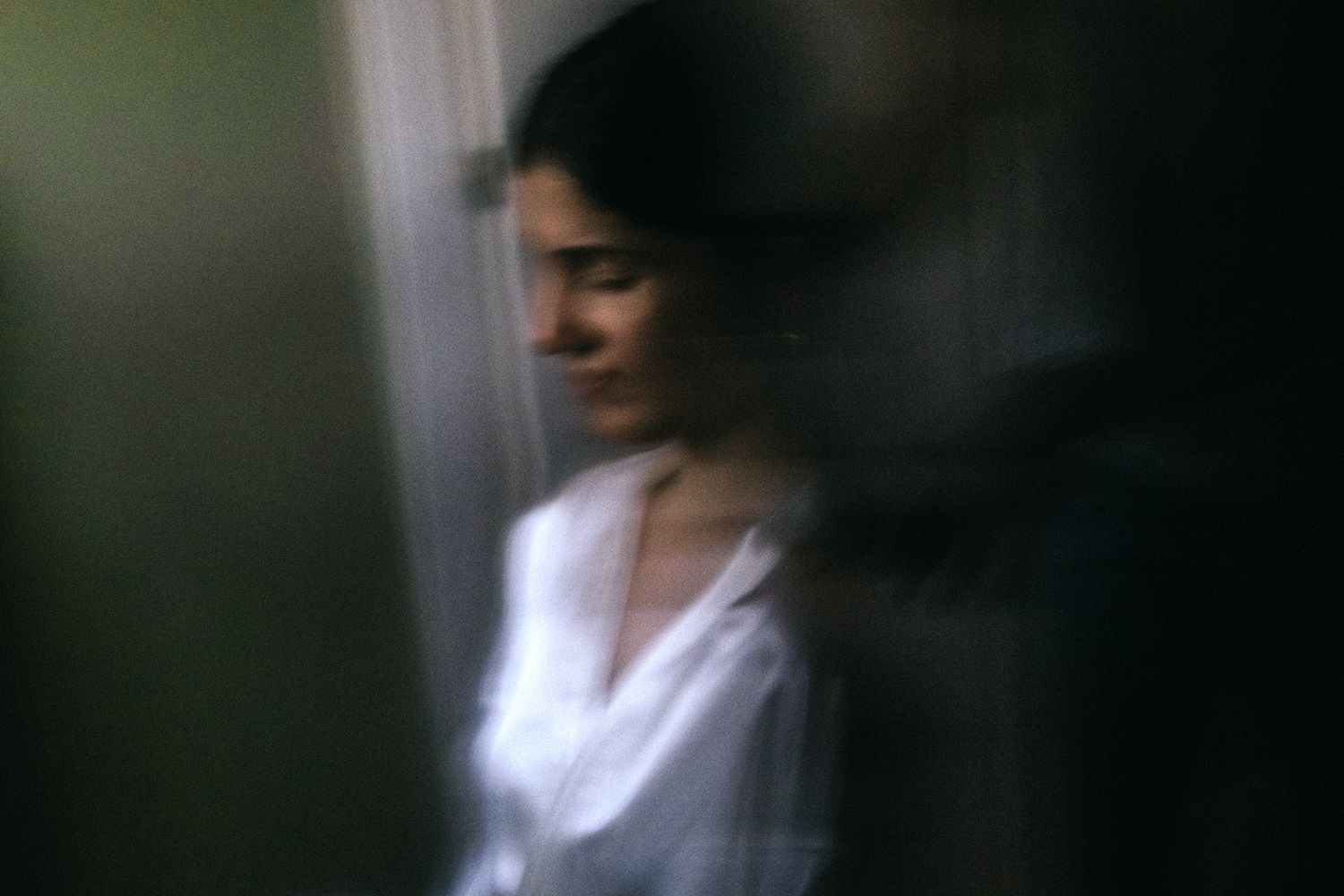 Blurred motion portrait of a woman in a white shirt standing near a window in soft, moody lighting.