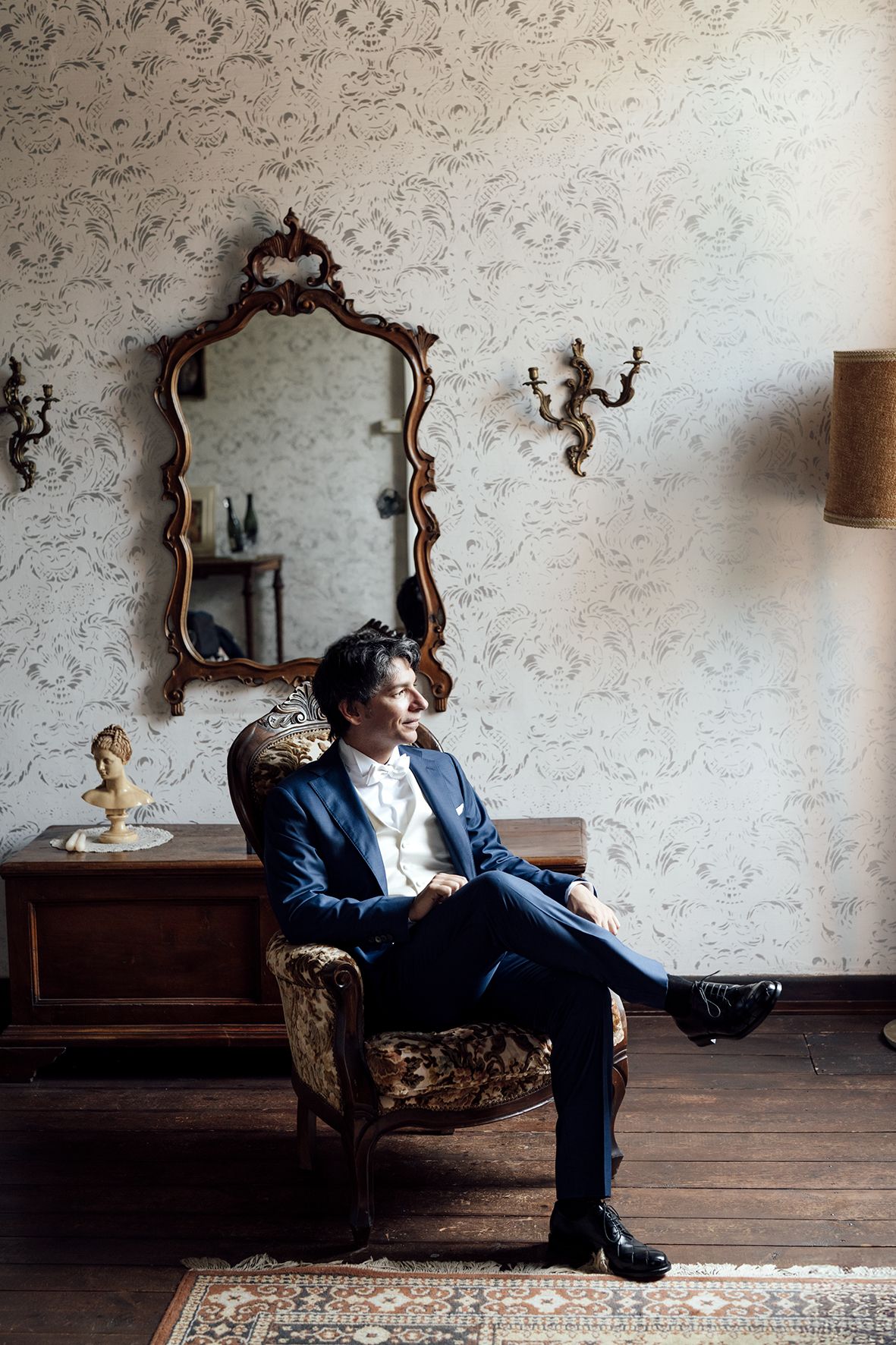 Person in formal attire relaxes on antique furniture against wallpapered wall with ornate mirror.