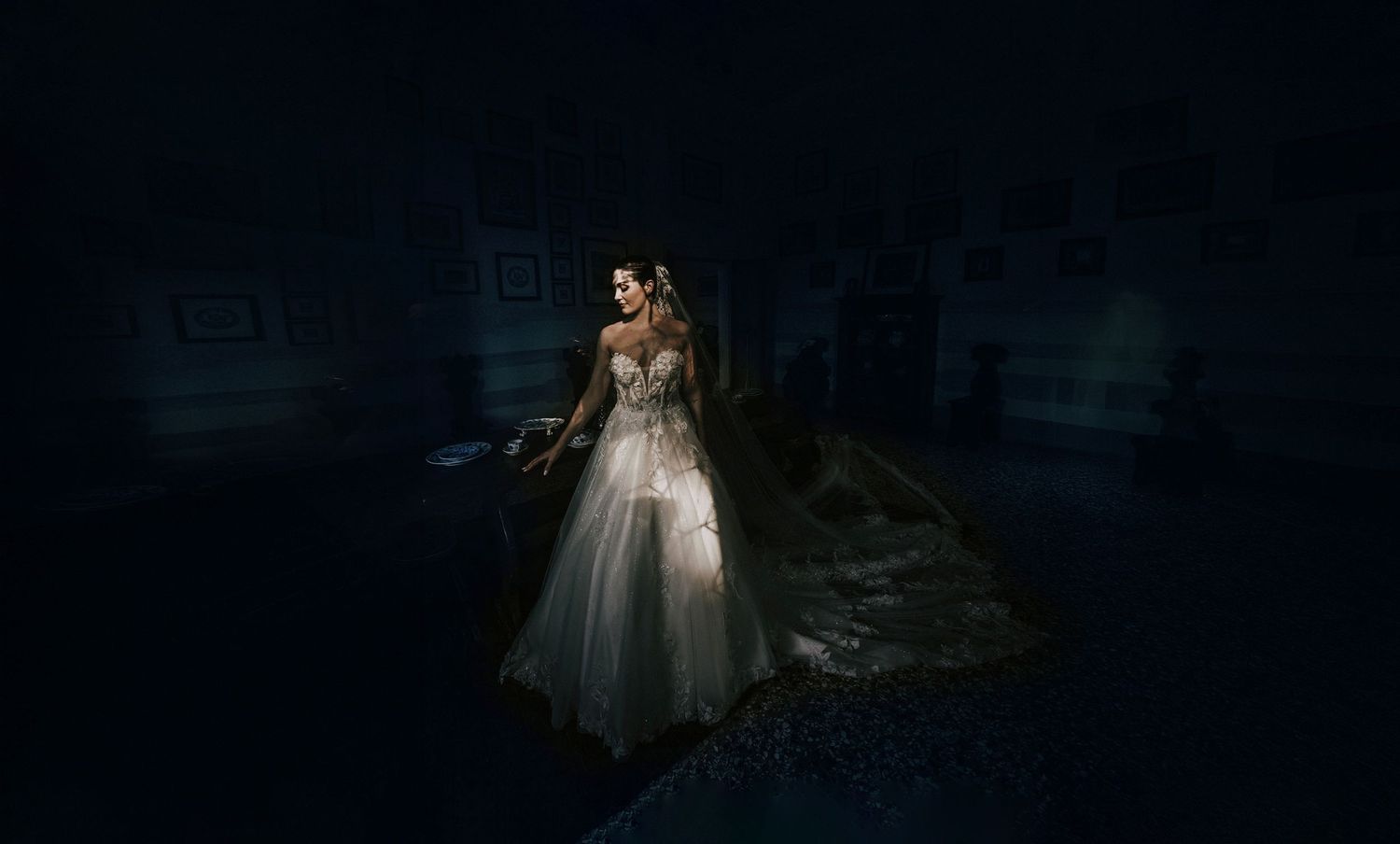 A bride in a white gown stands dramatically in a dark, moody room with dim lighting highlighting her elegant dress.