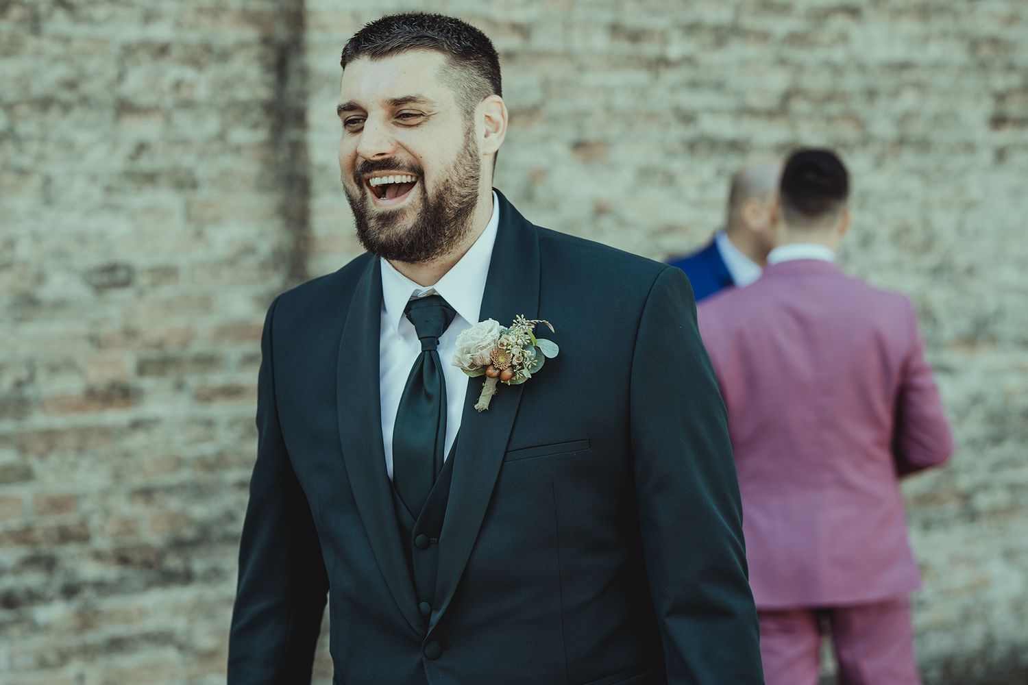 Wedding party members in formal attire stand against a rustic brick wall.