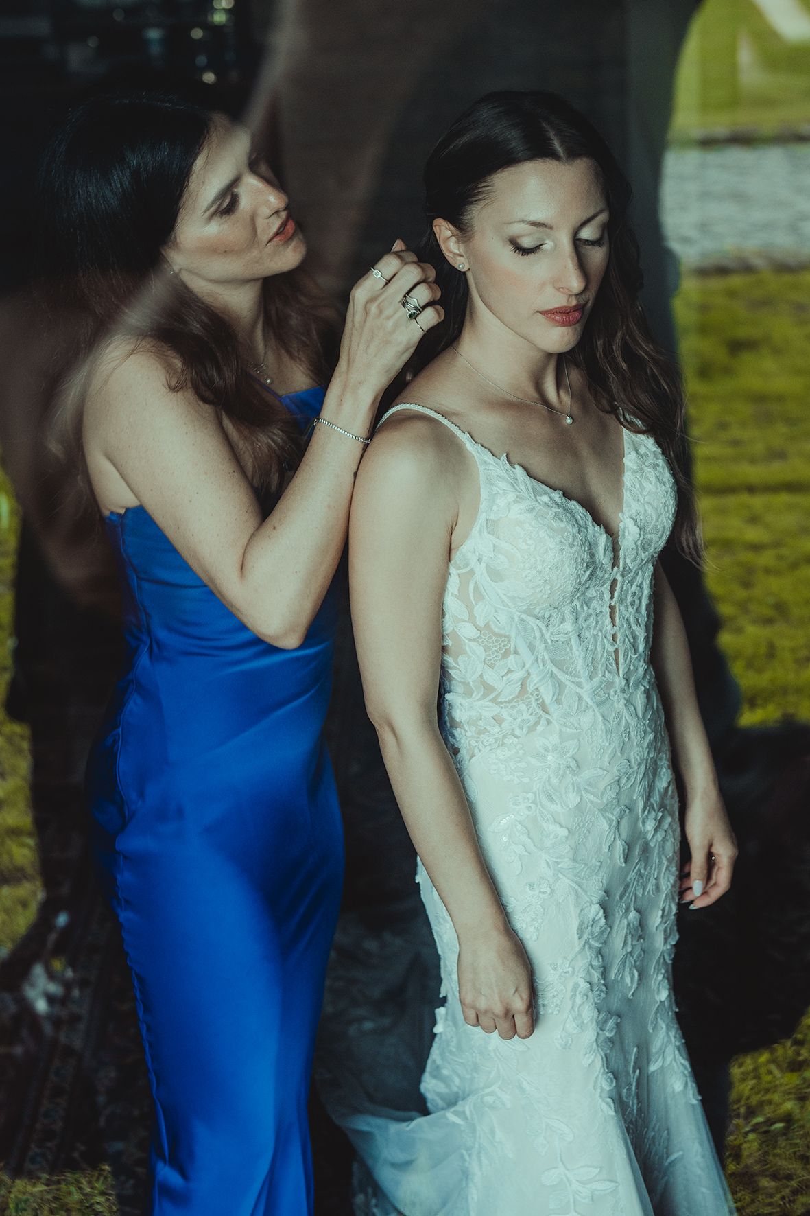 Two women in elegant dresses, one in royal blue and one in white lace, share a tender moment at a formal event.
