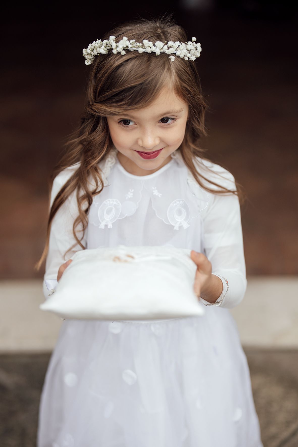 Flower girl in white dress wearing delicate floral crown holds ring pillow.