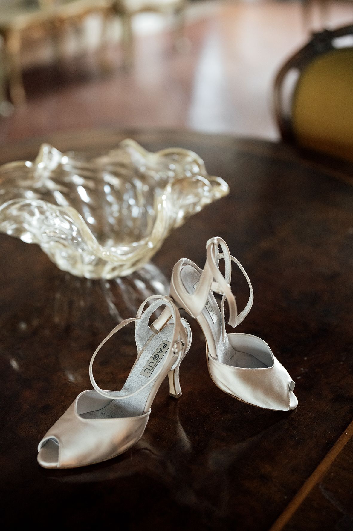 Elegant white bridal shoes displayed next to crystal bowl on wooden surface.