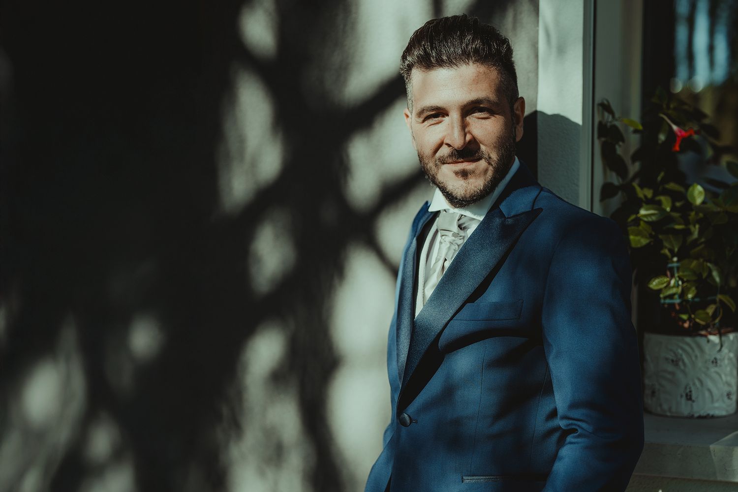 A groom in a navy blue suit smiles in natural lighting against a wall with shadows.