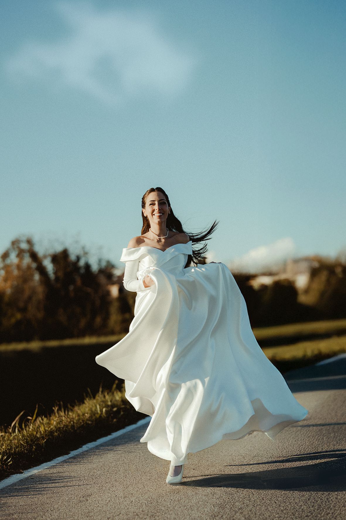 White wedding dress flowing dramatically against blue sky on outdoor path.