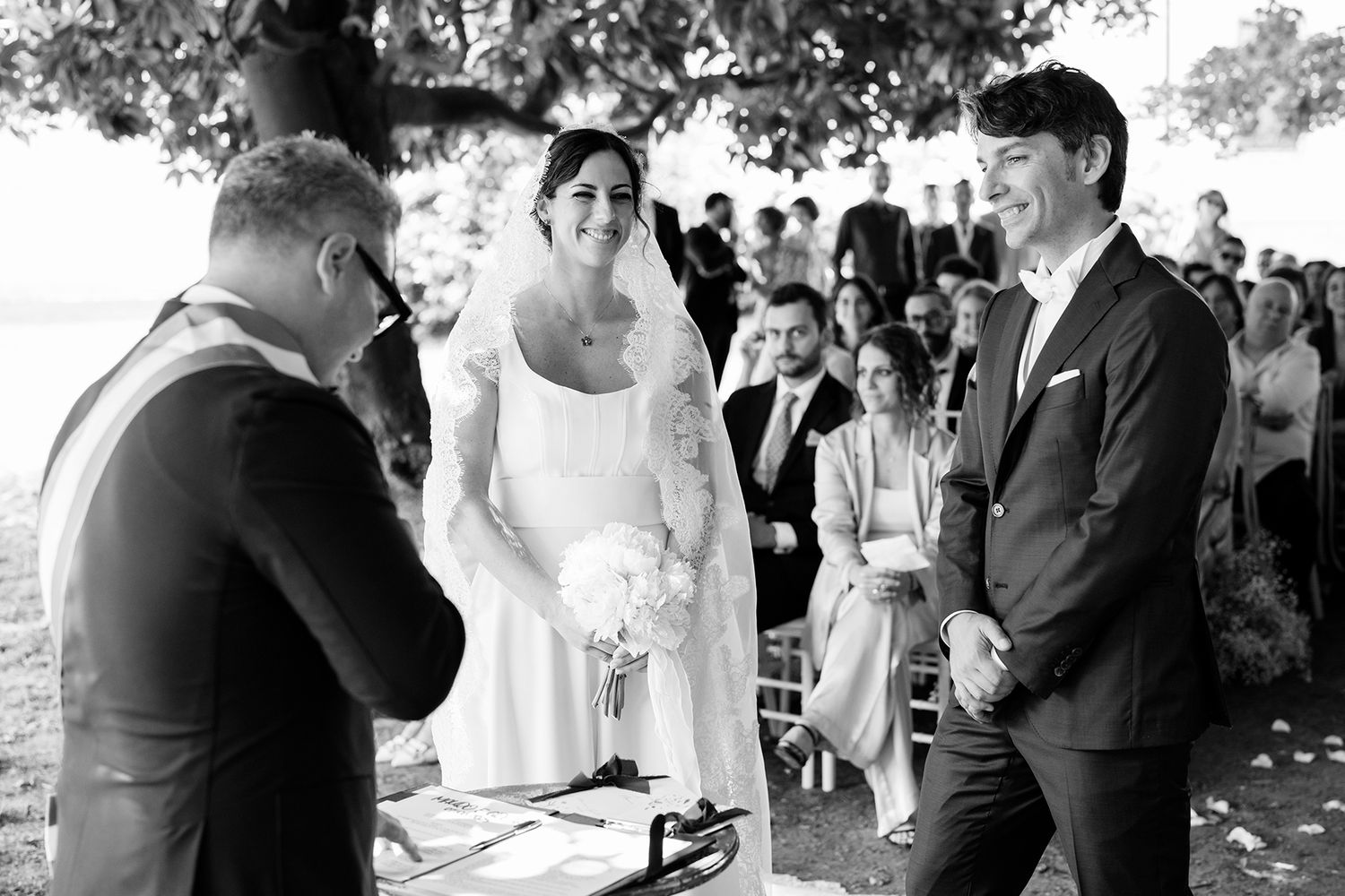 Wedding guests gathered outdoors under trees during a celebration.
