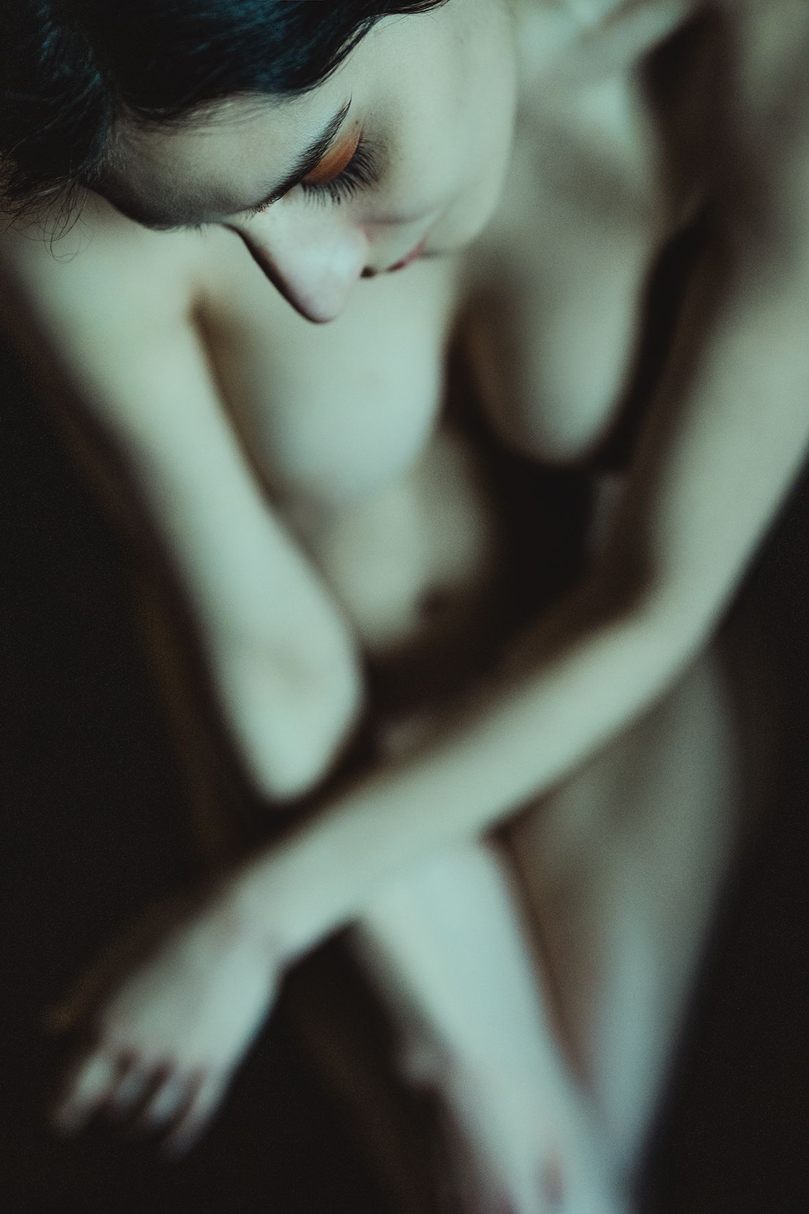 A woman with dark hair sits in a contemplative pose, arms crossed, in a moody, dimly lit artistic portrait.