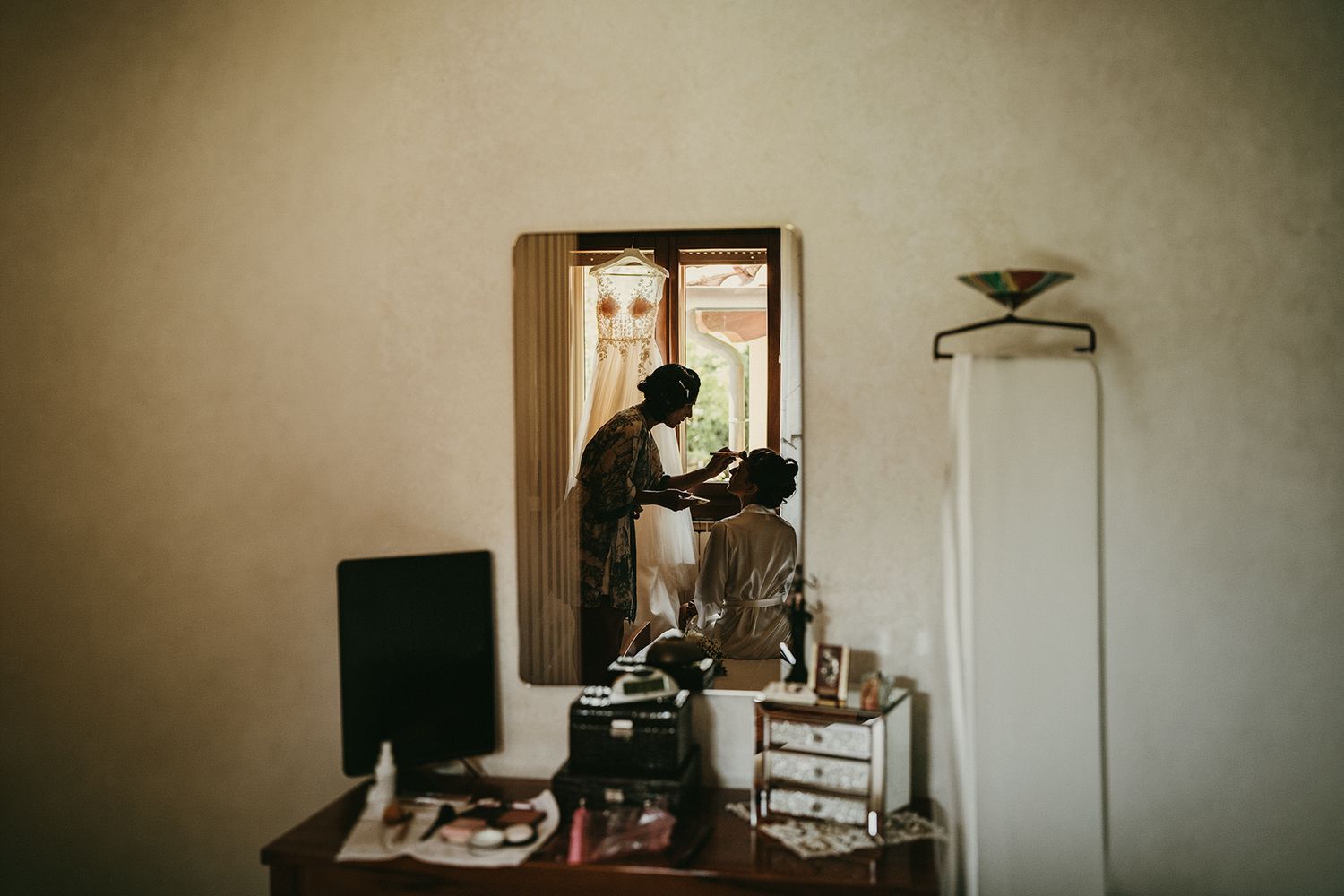 A hairstylist silhouetted by window light styles a bride's hair in a warmly lit rustic dressing room.