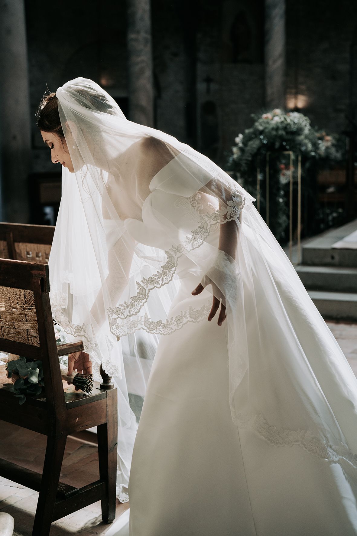 Elegant bride in white lace gown and flowing veil adjusts her dress in a church setting.
