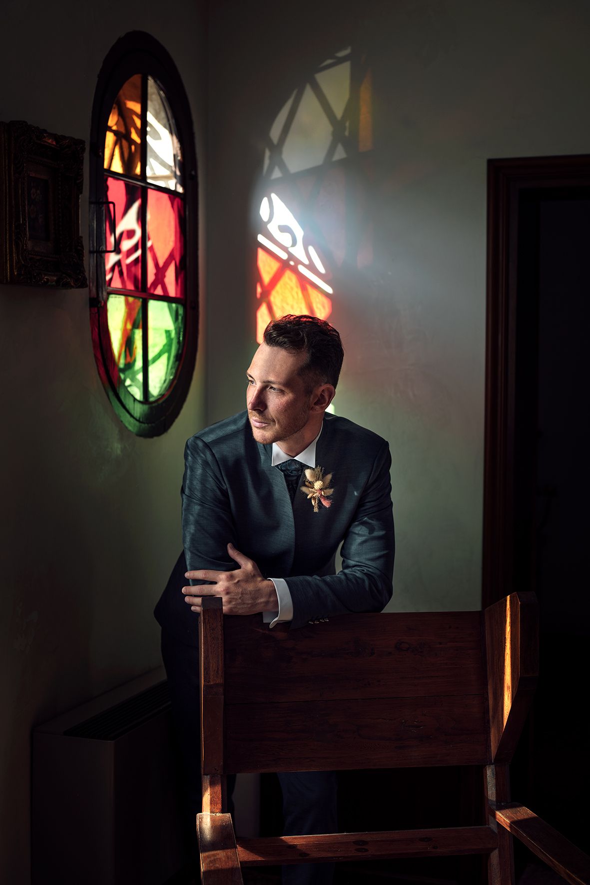 Dramatic portrait in church with stained glass window casting colorful light on suited figure.