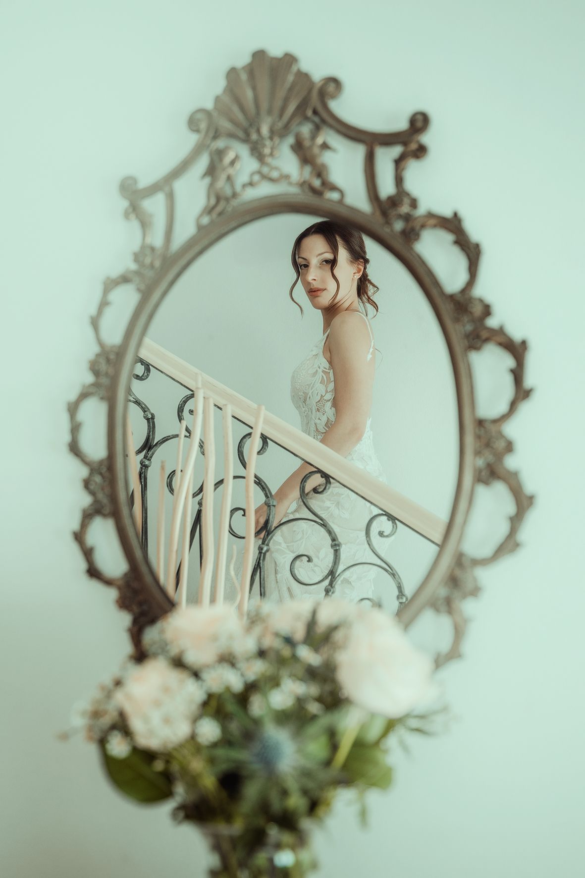 Artistic bridal portrait through ornate vintage mirror frame with flowers in foreground.