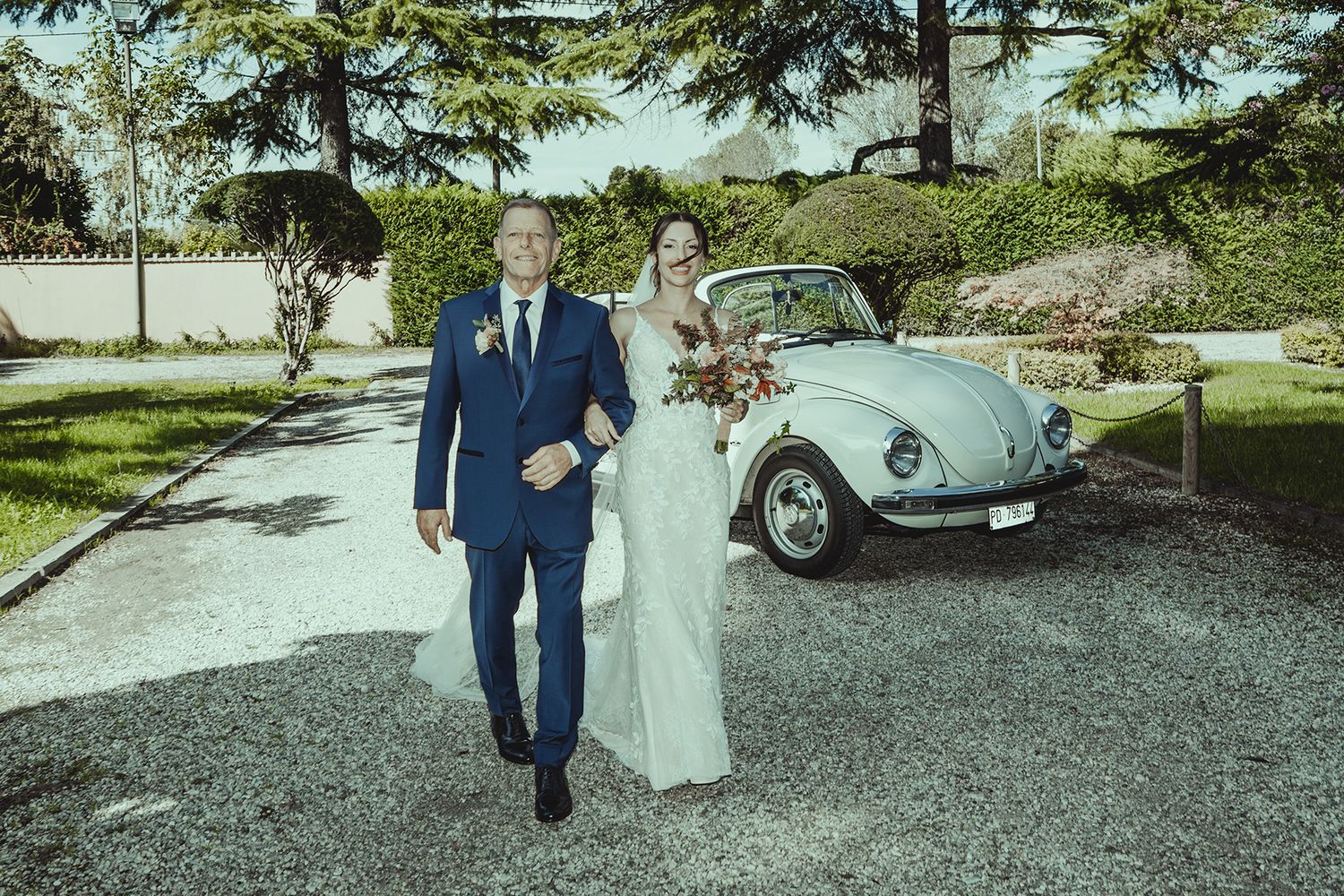 Classic Volkswagen Beetle parked on gravel near trees at outdoor wedding venue.