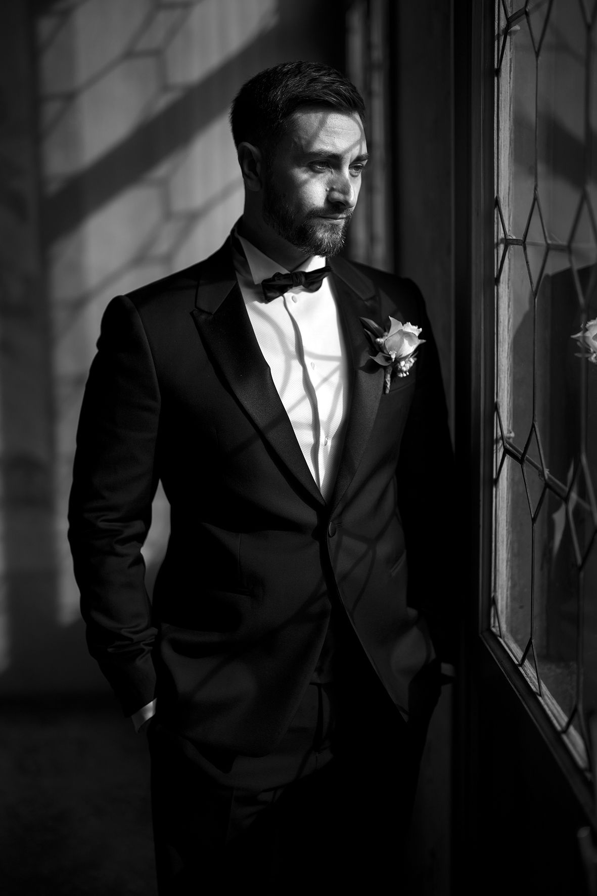 Dramatic black and white portrait of a groom in a tuxedo standing by a window with moody lighting.