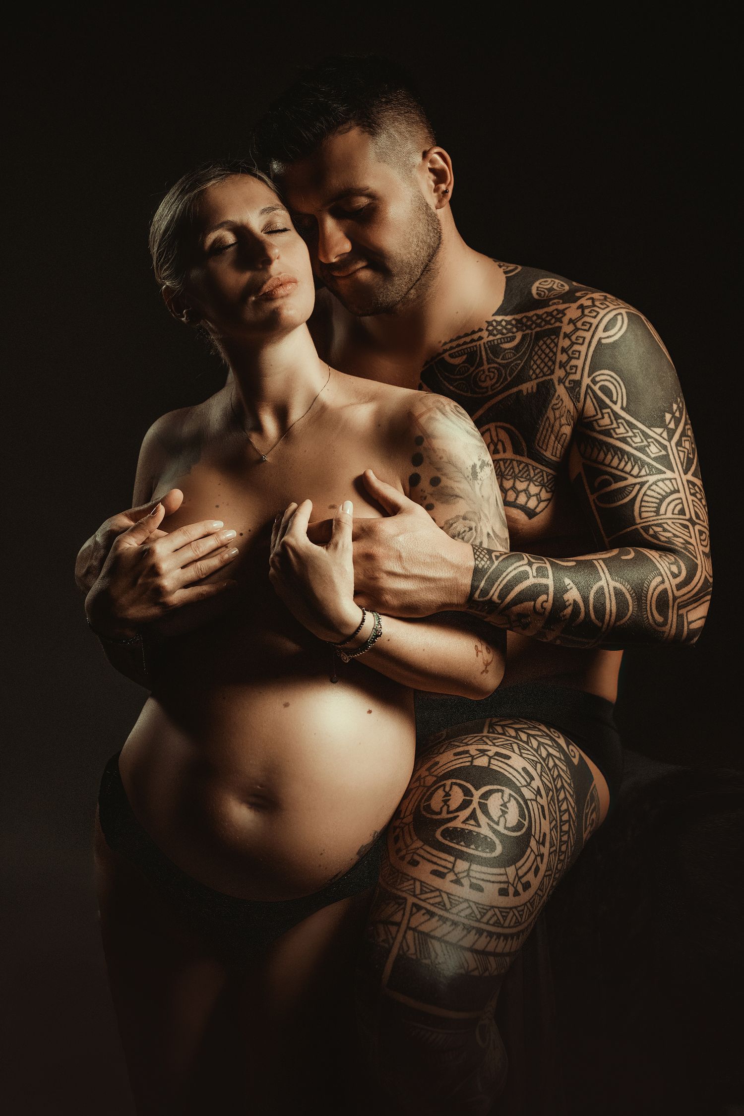 Artistic maternity photo featuring intimate couple pose with tribal tattoos in dark lighting.