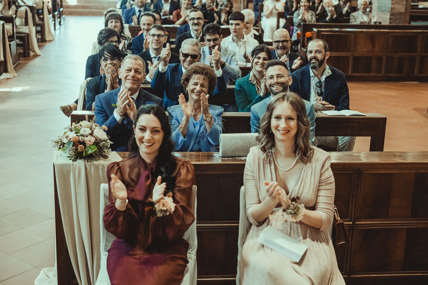 Wedding guests smile and applaud while seated in wooden church pews.