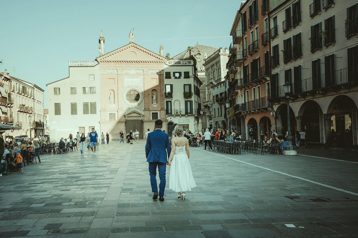 A couple in wedding attire walks down a historic European street with classic architecture.