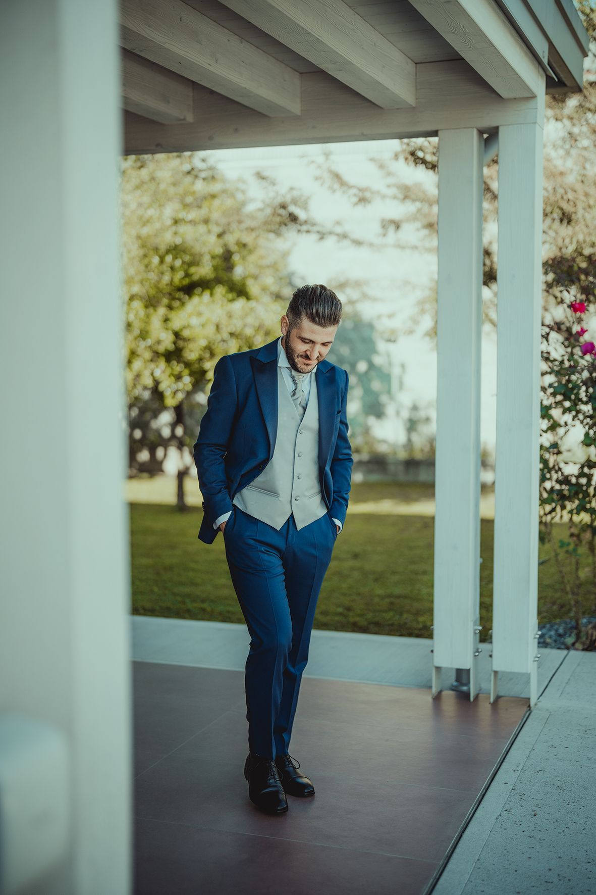 A stylish suit-clad figure poses on a covered porch with autumn scenery in background.