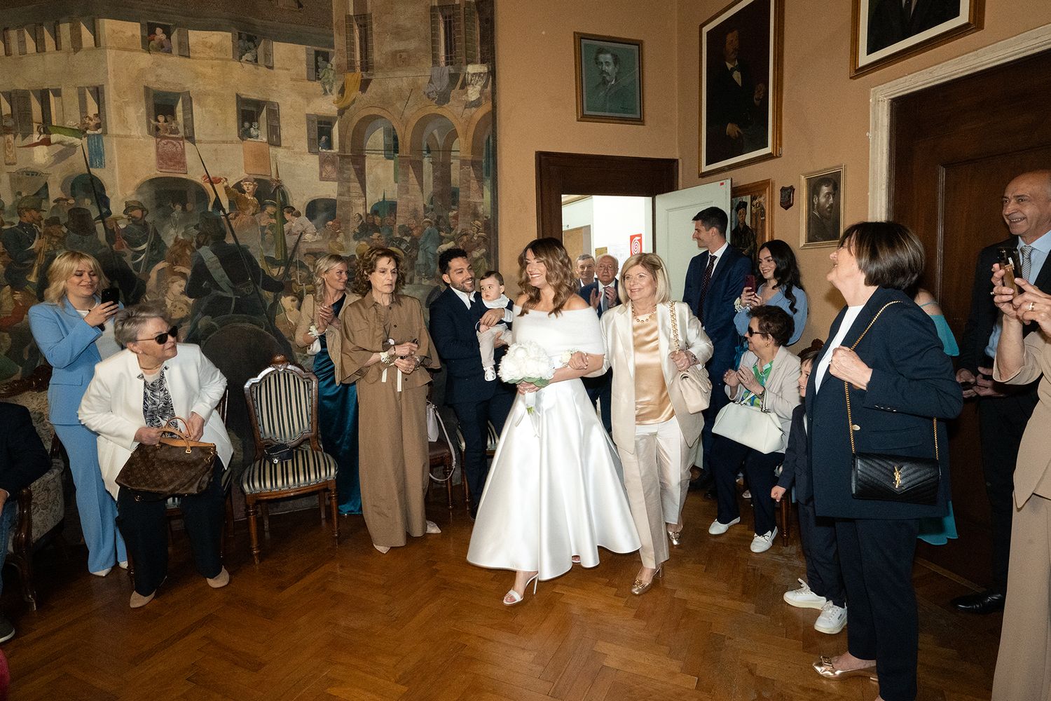 Wedding guests gathered in an ornate room with hardwood floors and decorative wall murals.