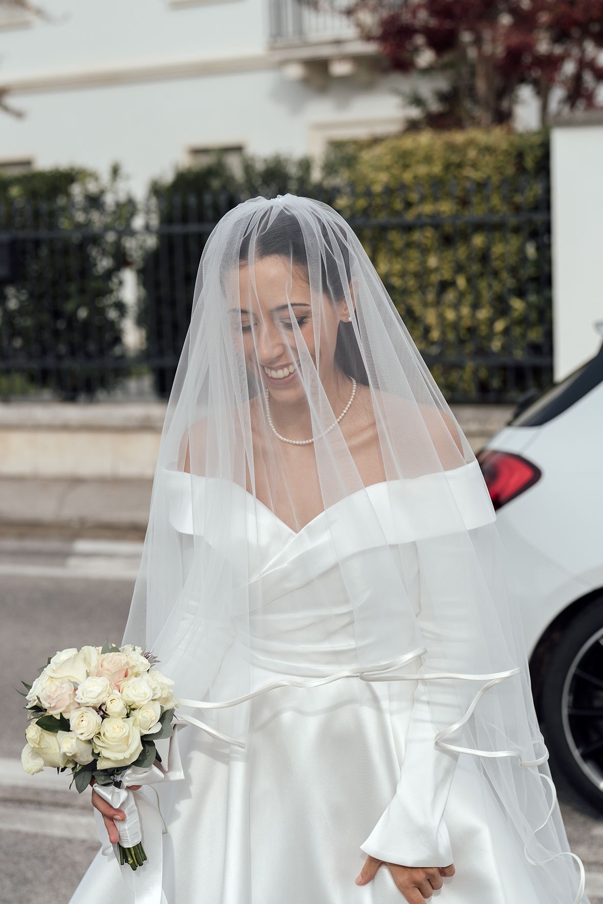 Joyful bride in a structured white dress with portrait collar and delicate veil holding white roses.