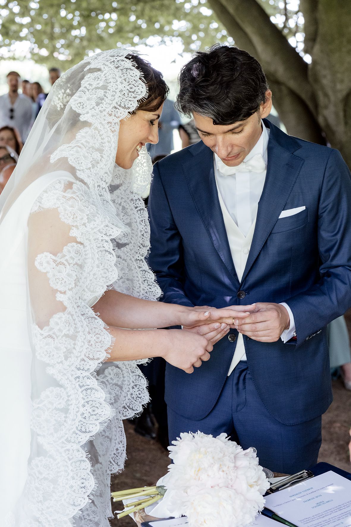 Couple in navy suit and lace veil examining wedding documents during outdoor ceremony.