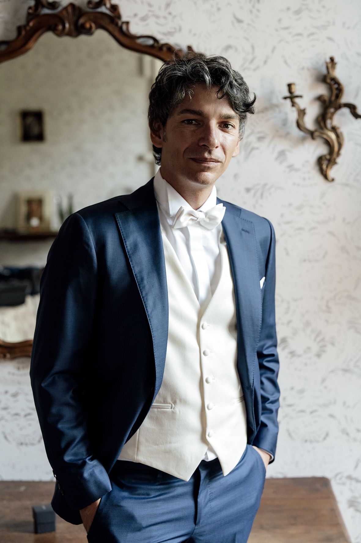 Person wearing navy blue suit and white dress shirt poses in an elegant interior setting.
