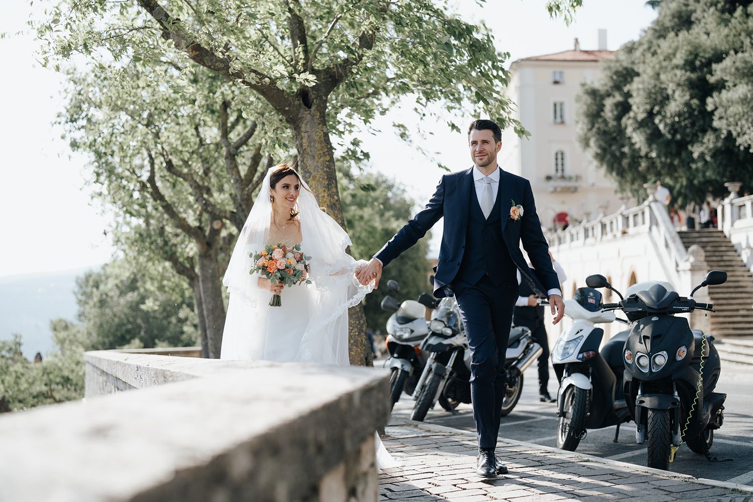 Wedding couple poses on scenic Mediterranean pathway with motorcycles in background.