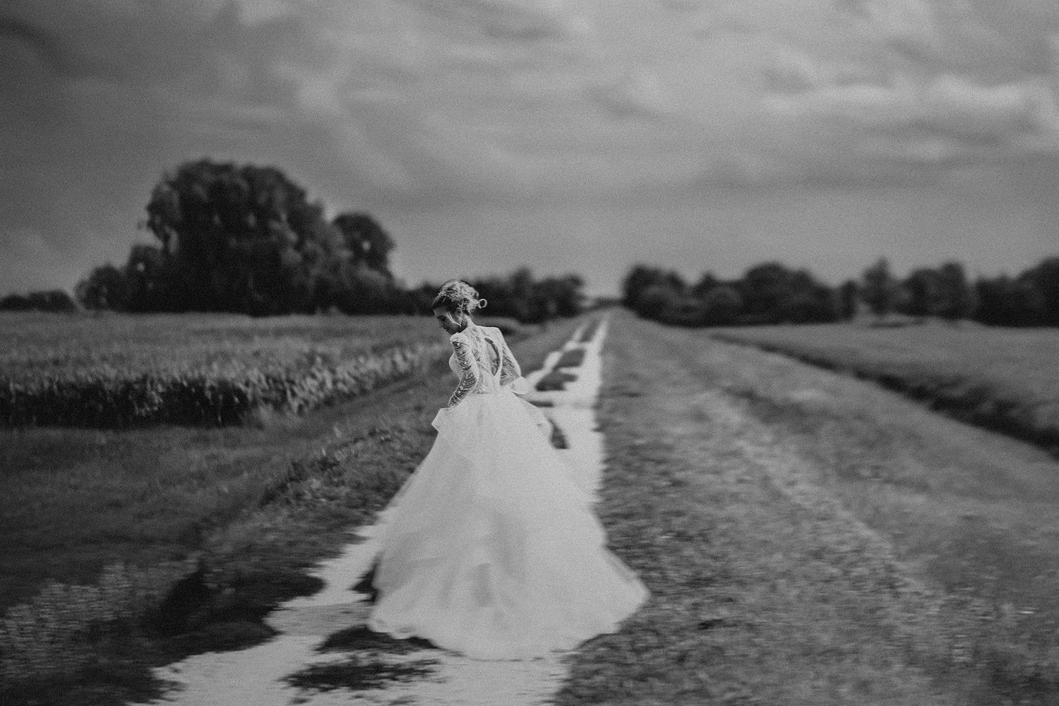 Bride in flowing white gown walks along a rural dirt path flanked by fields, captured in moody black and white.