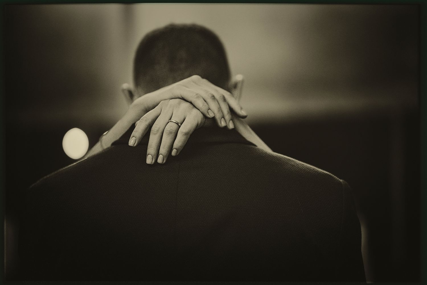 Black and white artistic photograph showing a hand resting on the back of someone's shoulder in dim lighting.