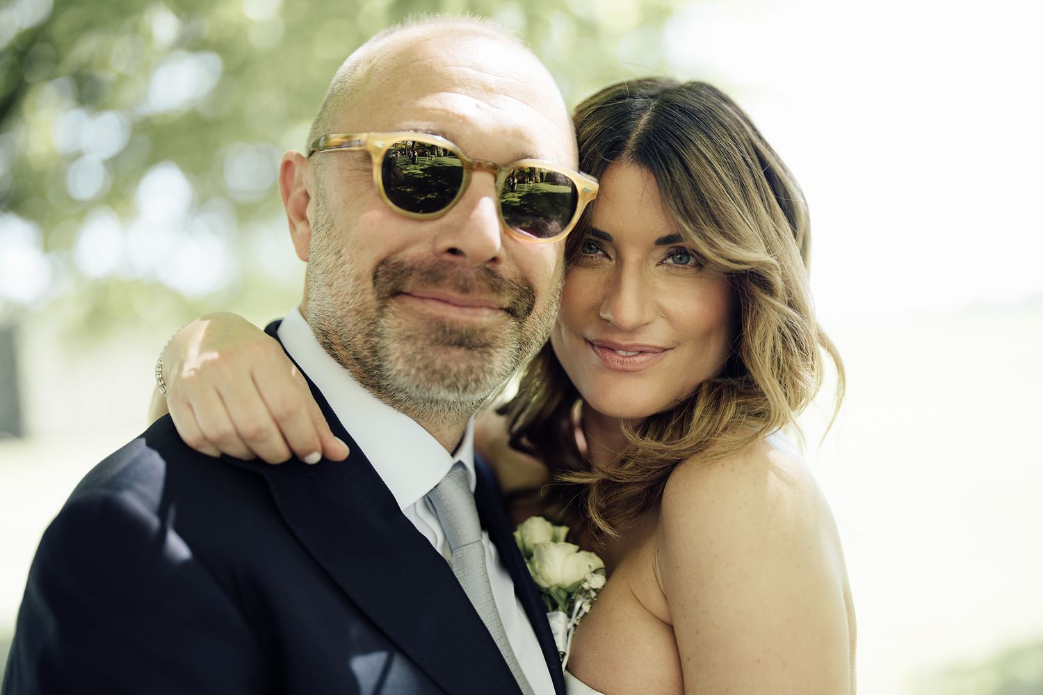 Wedding couple poses together outdoors in formal attire with sunglasses and smiles.