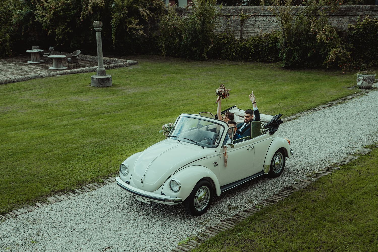 Mint green vintage Volkswagen Beetle convertible drives along garden path after wedding ceremony.