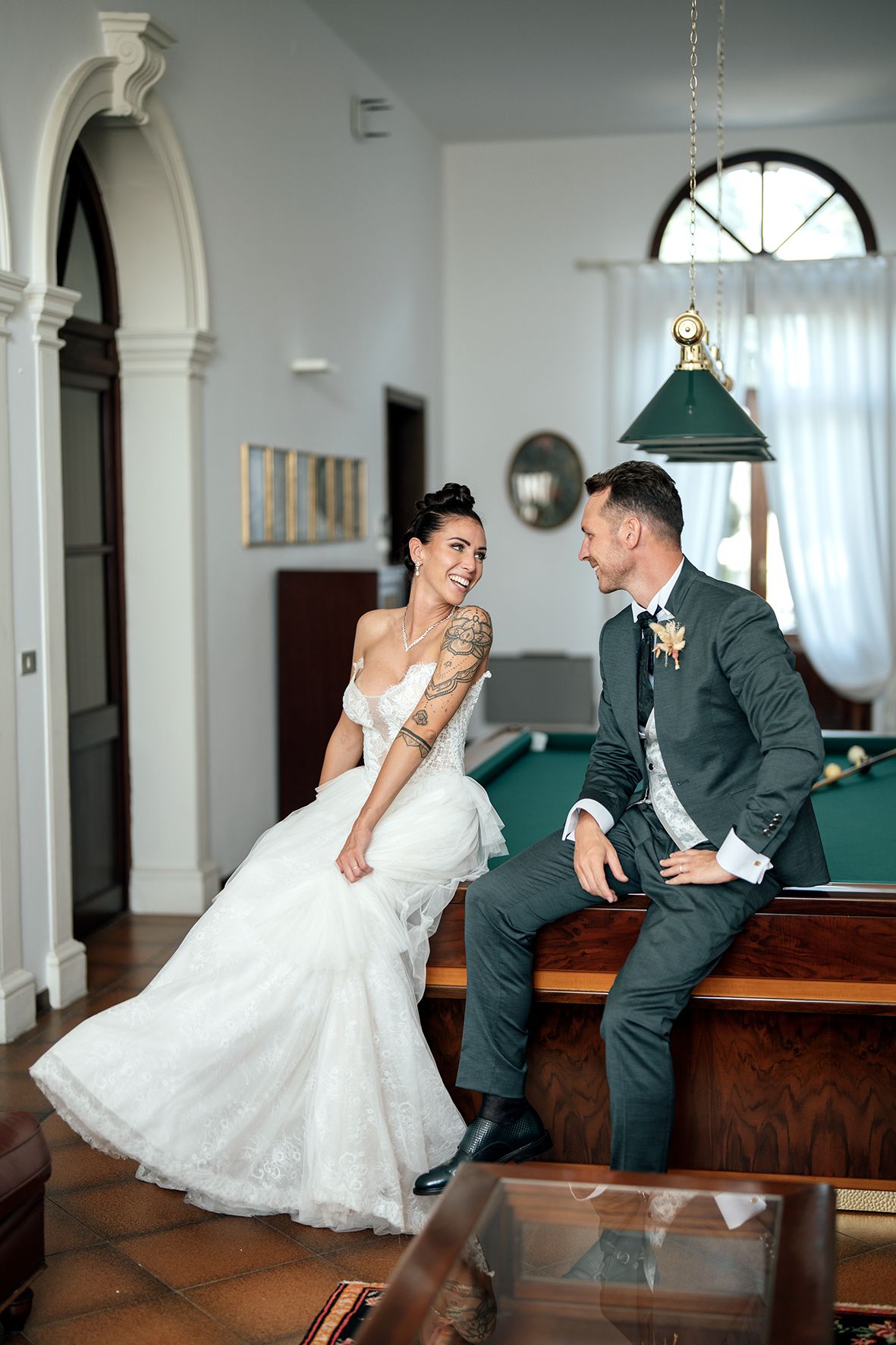 Playful wedding couple poses together on vintage pool table in elegant indoor setting.