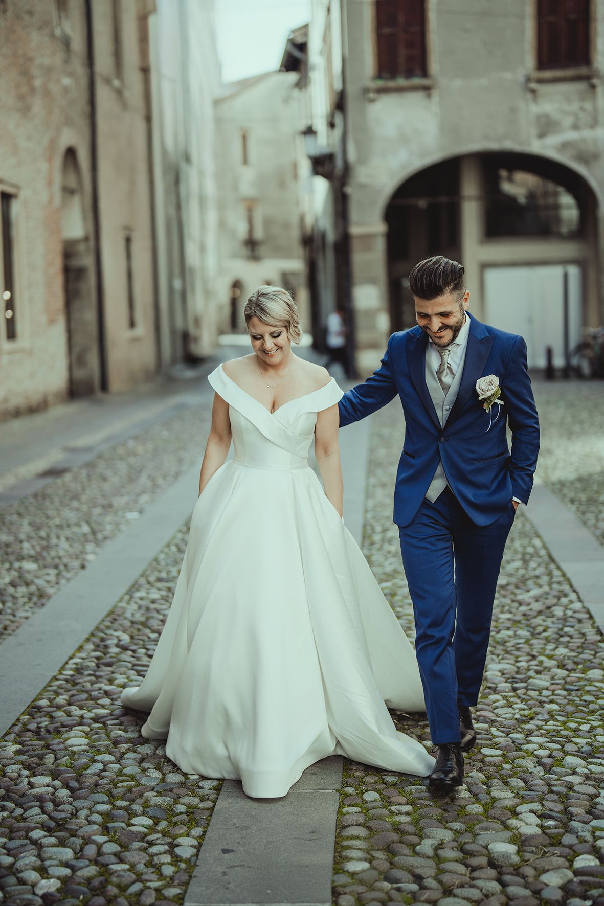 Newlyweds in formal attire walking together on a cobblestone street in an old European town.