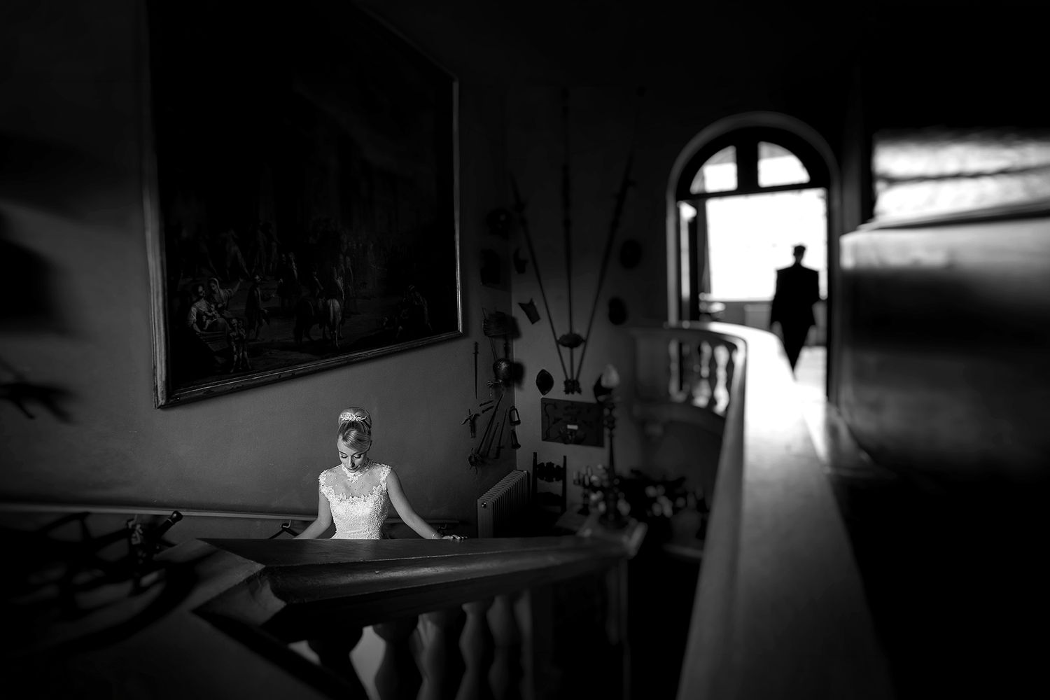 Black and white wedding photo of bride at staircase railing with groom silhouette in arched doorway.