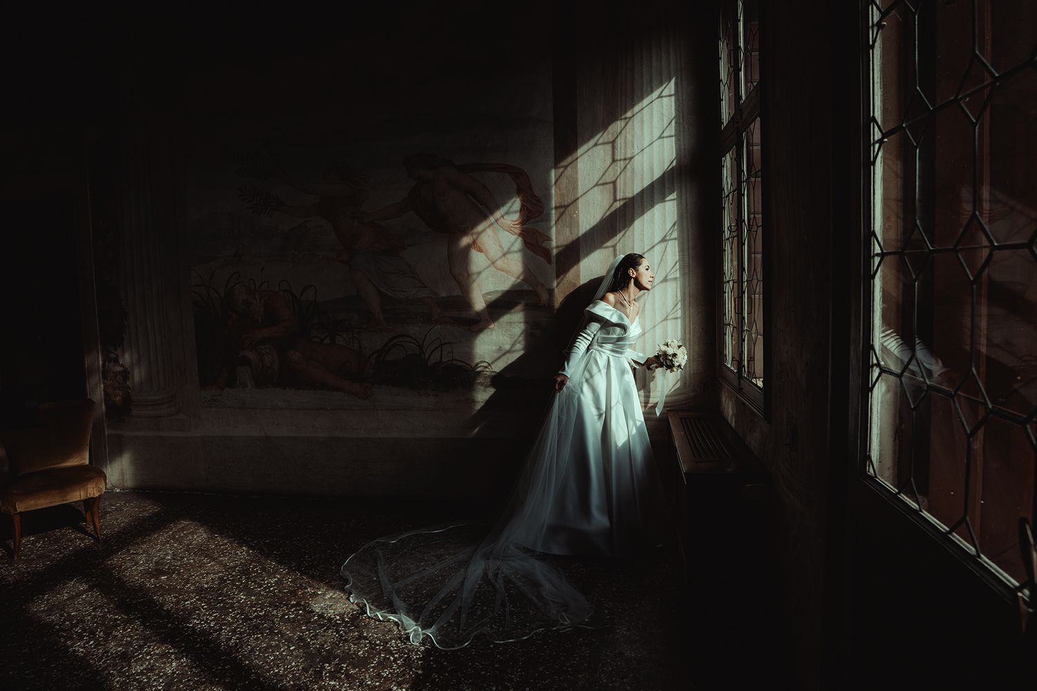 Artistic silhouette of a bride in flowing white gown standing by a dramatic window in dark setting.