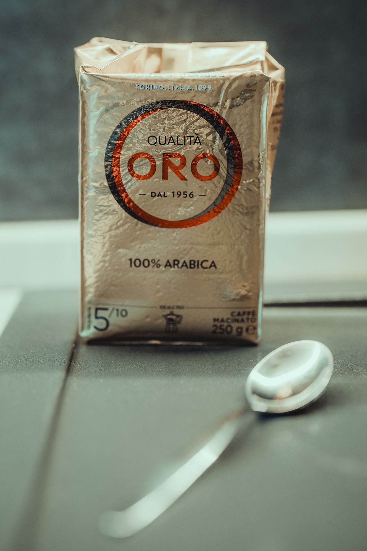 Package of ORO premium 100% Arabica coffee with white spoon on gray surface.