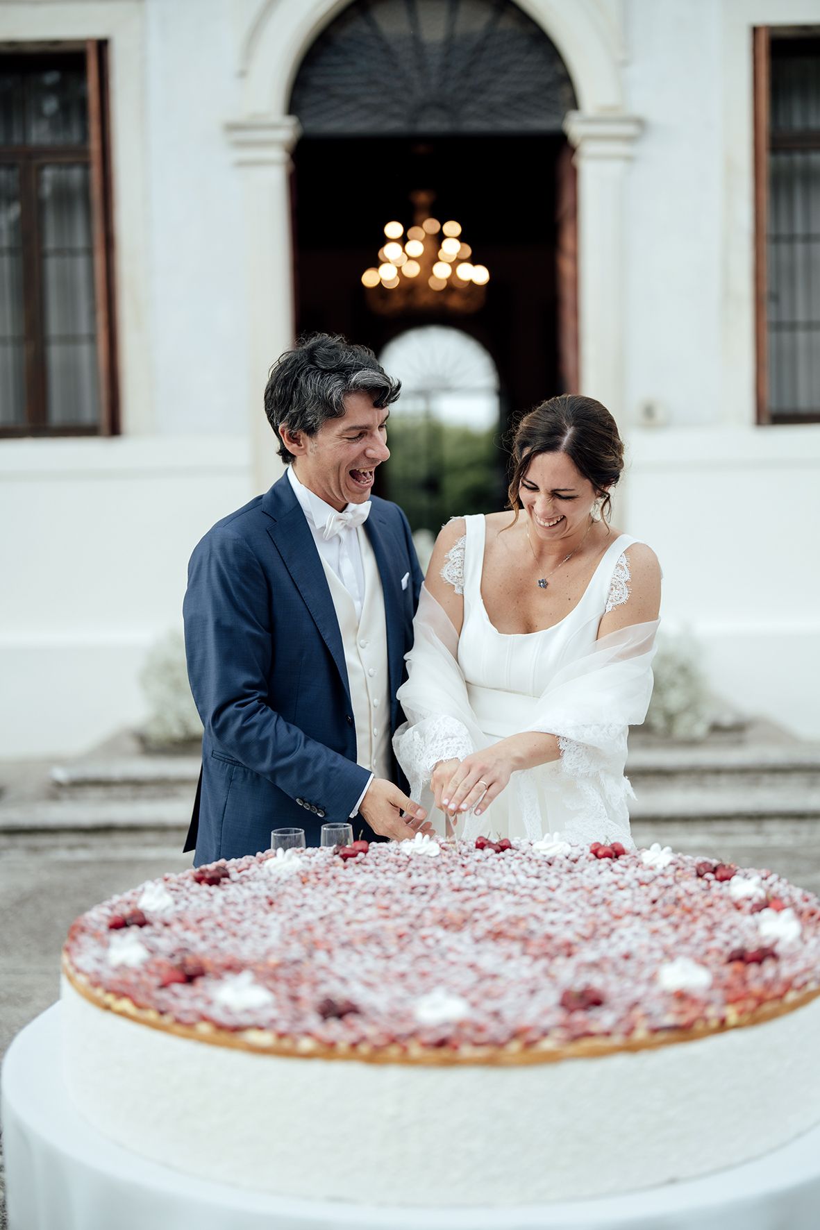 Couple cutting their wedding cake decorated with scattered rose petals on a white table.