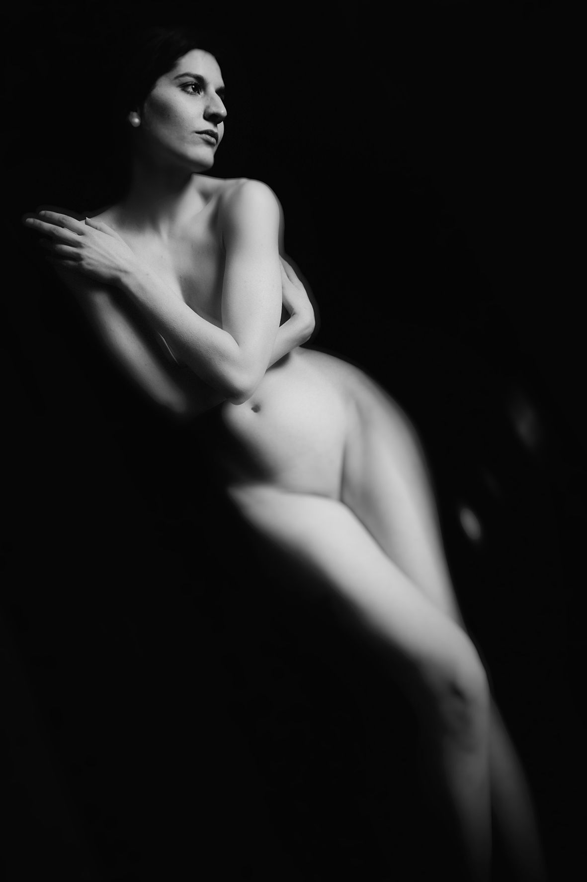 Black and white fine art nude photograph of a woman seated in dramatic chiaroscuro lighting against a dark background.