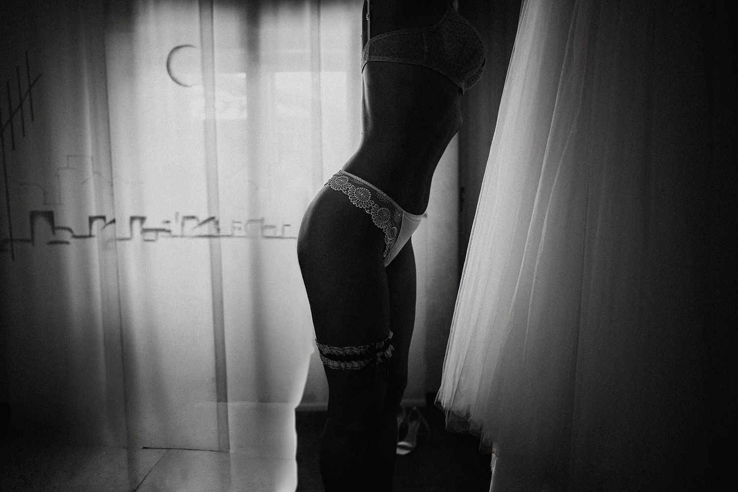 Black and white photo of a woman in lingerie standing by sheer curtains in dramatic window light.